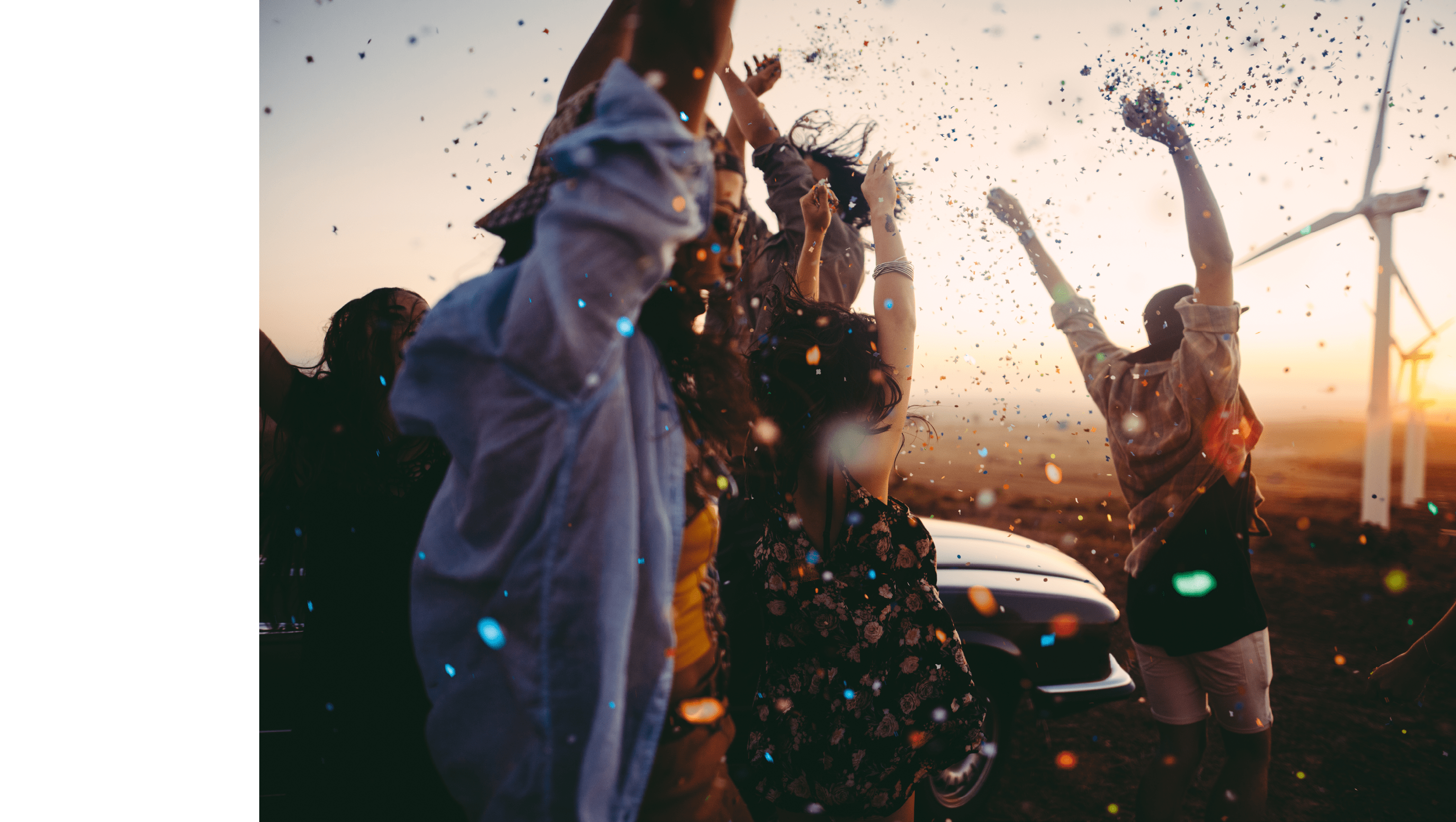 Group of multi-ethnic teens dressed in boho style partying outdoors throwing colourful confetti in a wind farm at sunset during their american carefree road trip