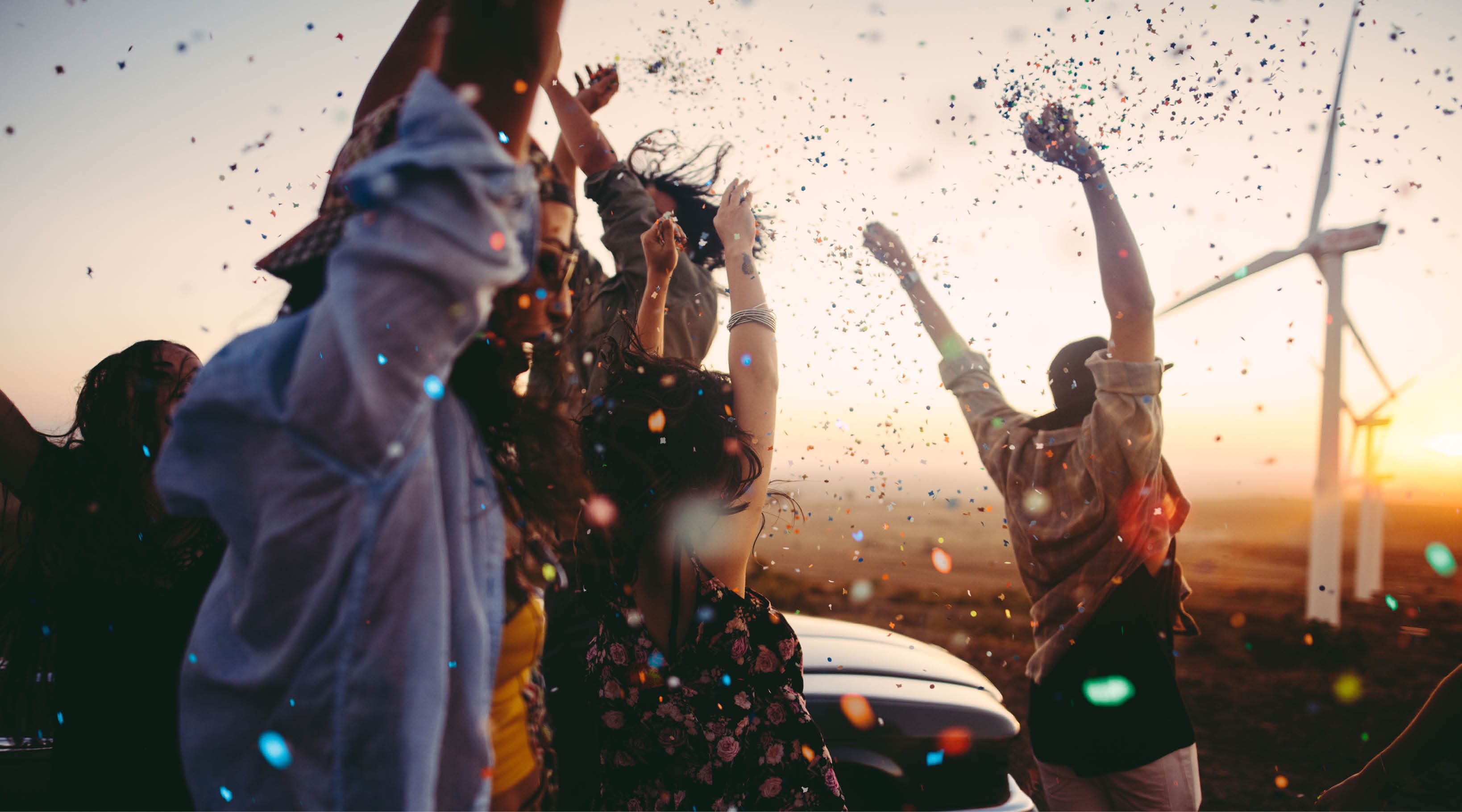 Group of multi-ethnic teens dressed in boho style partying outdoors throwing colourful confetti in a wind farm at sunset during their american carefree road trip