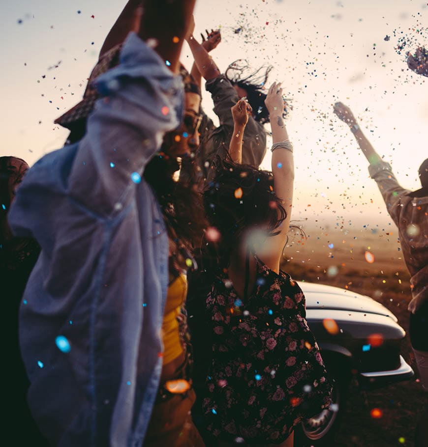 Group of multi-ethnic teens dressed in boho style partying outdoors throwing colourful confetti in a wind farm at sunset during their american carefree road trip